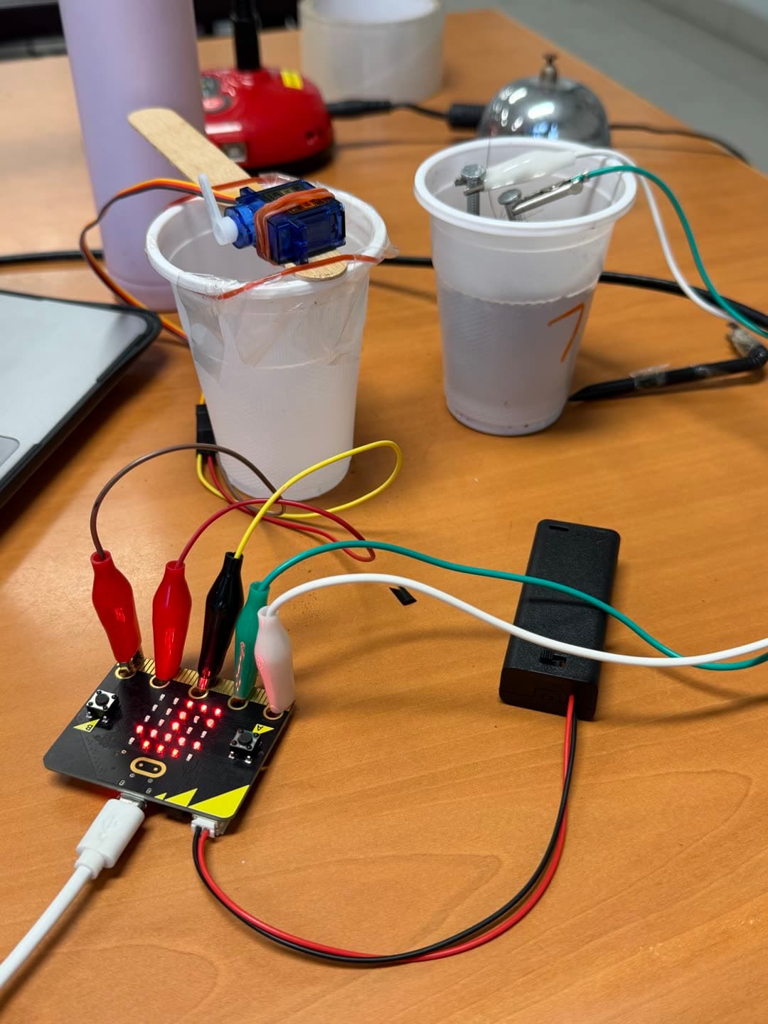 Close-up: Micro:bit lit up, alligator clips, and cup-based watering mechanism with servo paddle